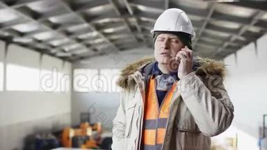 一个戴着安全帽、穿着工作服的人正站在机库的中央，对着镜头用手机说话